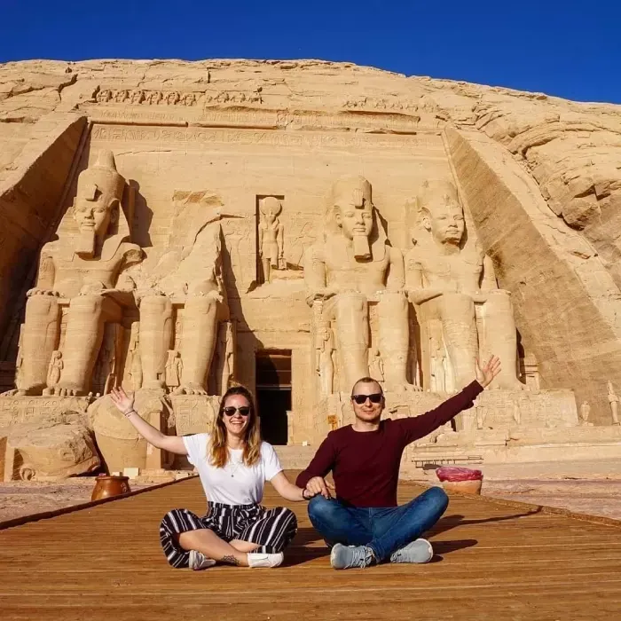  two visitors sit on a wooden platform in front of the Ancient Temple of Abu Simbel with seated statues of Pharaoh Ramesses II, beneath a clear blue sky.