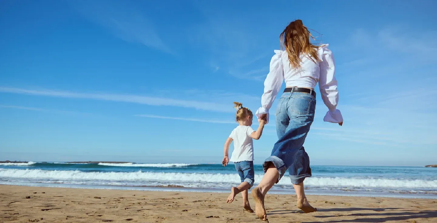 Joven madre y su pequeña hija caminando descalzas de la mano por la playa de arena.