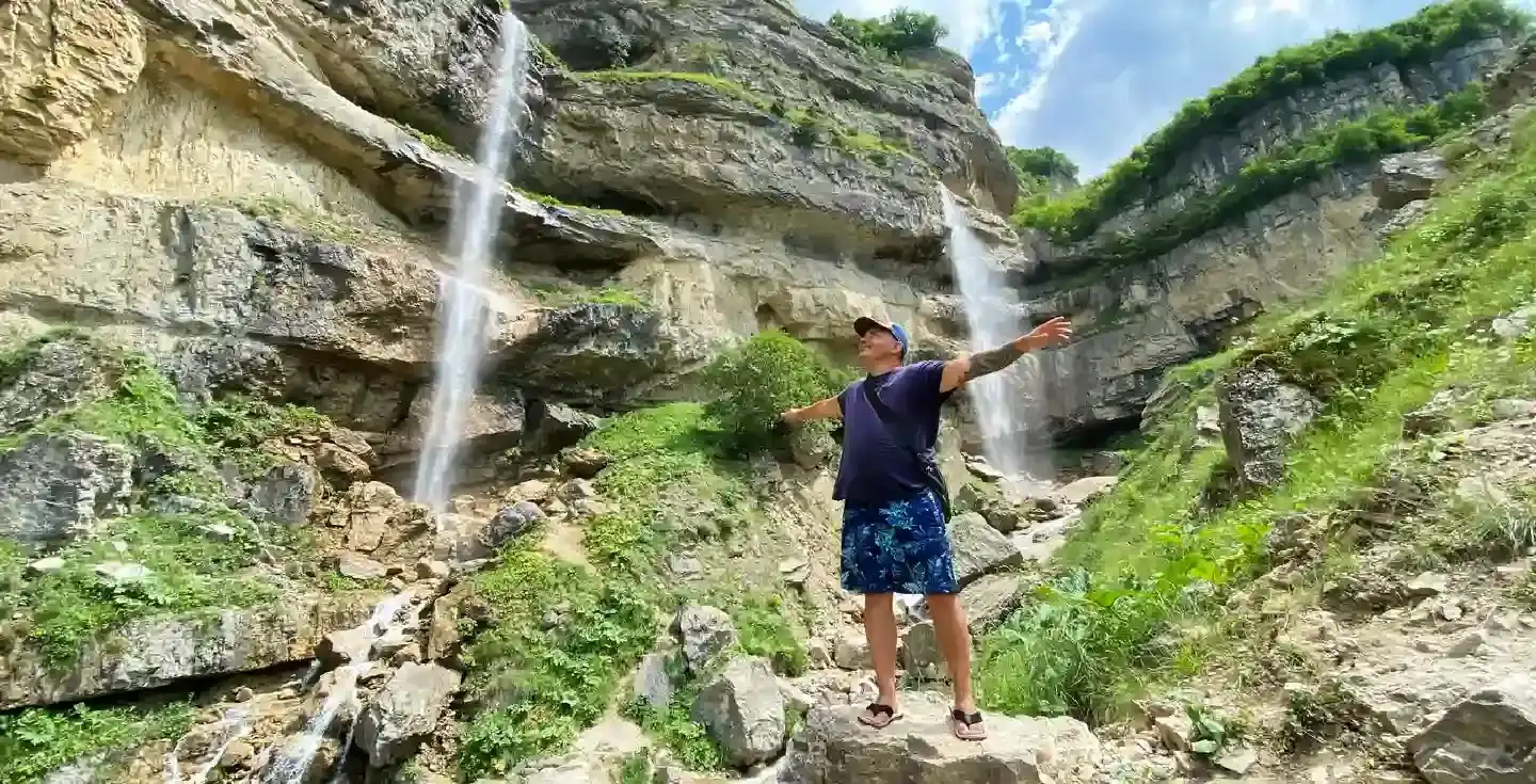 Happy guy stands on a cliff in front of two amazing waterfalls, arms outstretched. Nature of Azerbaijan. Adventures and tourism in the village of Laza. Scene is serene and peaceful.