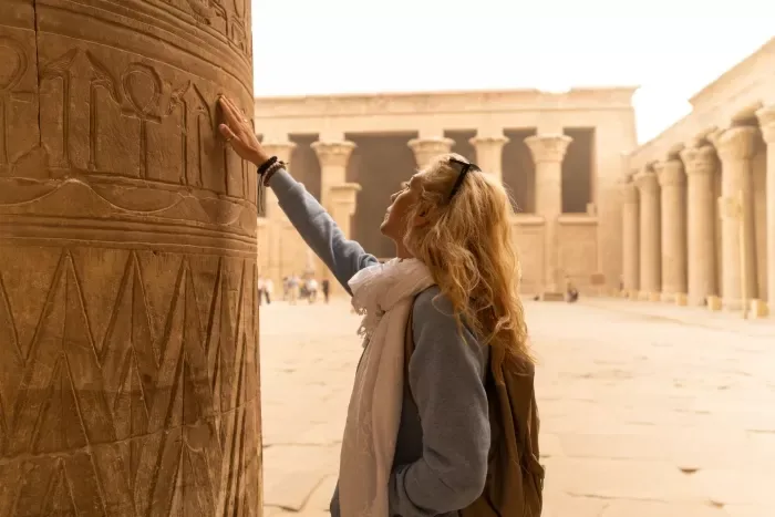 a blonde woman in gray shert and white scarf  touching ancient column at Egyptian temple ruins.