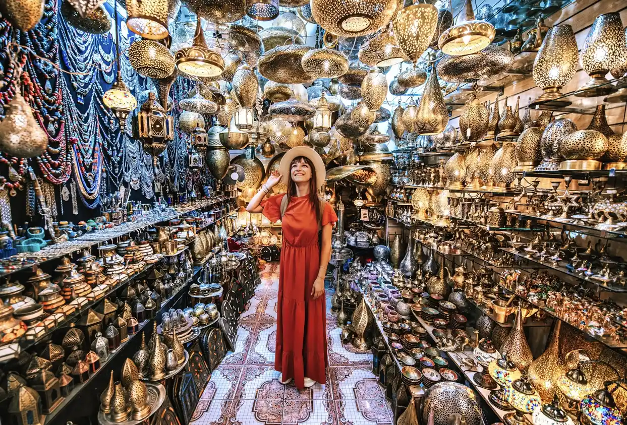 A woman in a red dress is in a Moroccan souk, surrounded by an abundance of ornate lamps and colorful jewelry.