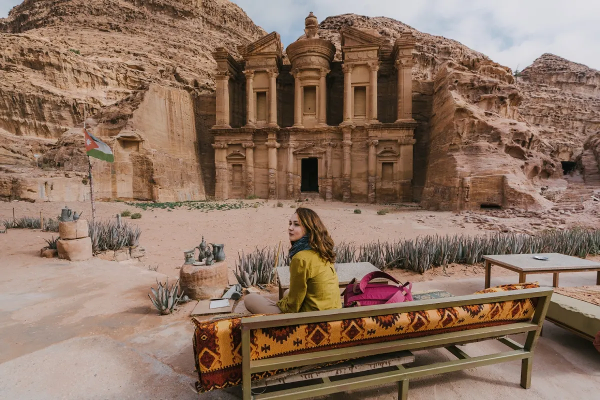 Person seated on patterned bench before Petra’s Monastery carved into sandstone cliffs with Jordanian flag nearby.