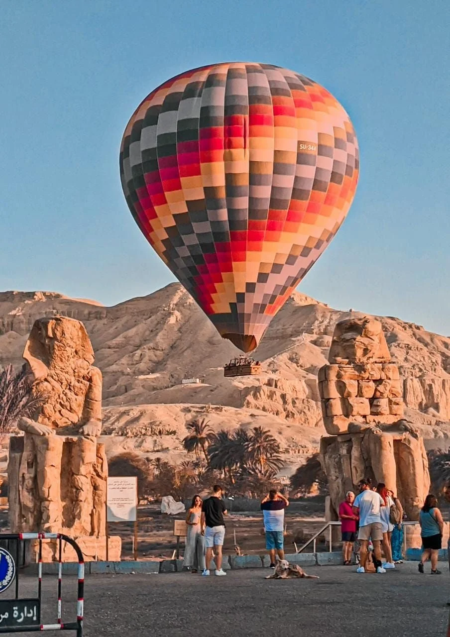 Hot air balloon floating between the Colossi of Memnon in Luxor, Egypt, with tourists observing the scene and desert mountains in the background during sunrise.