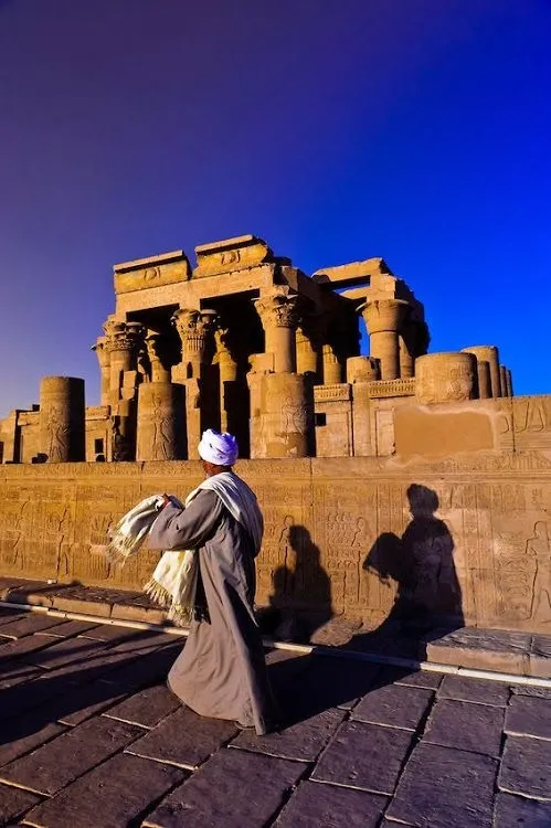 A man in traditional Egyptian clothing walking past the ancient Kom Ombo Temple at sunset, with carved hieroglyphics and columns in the background