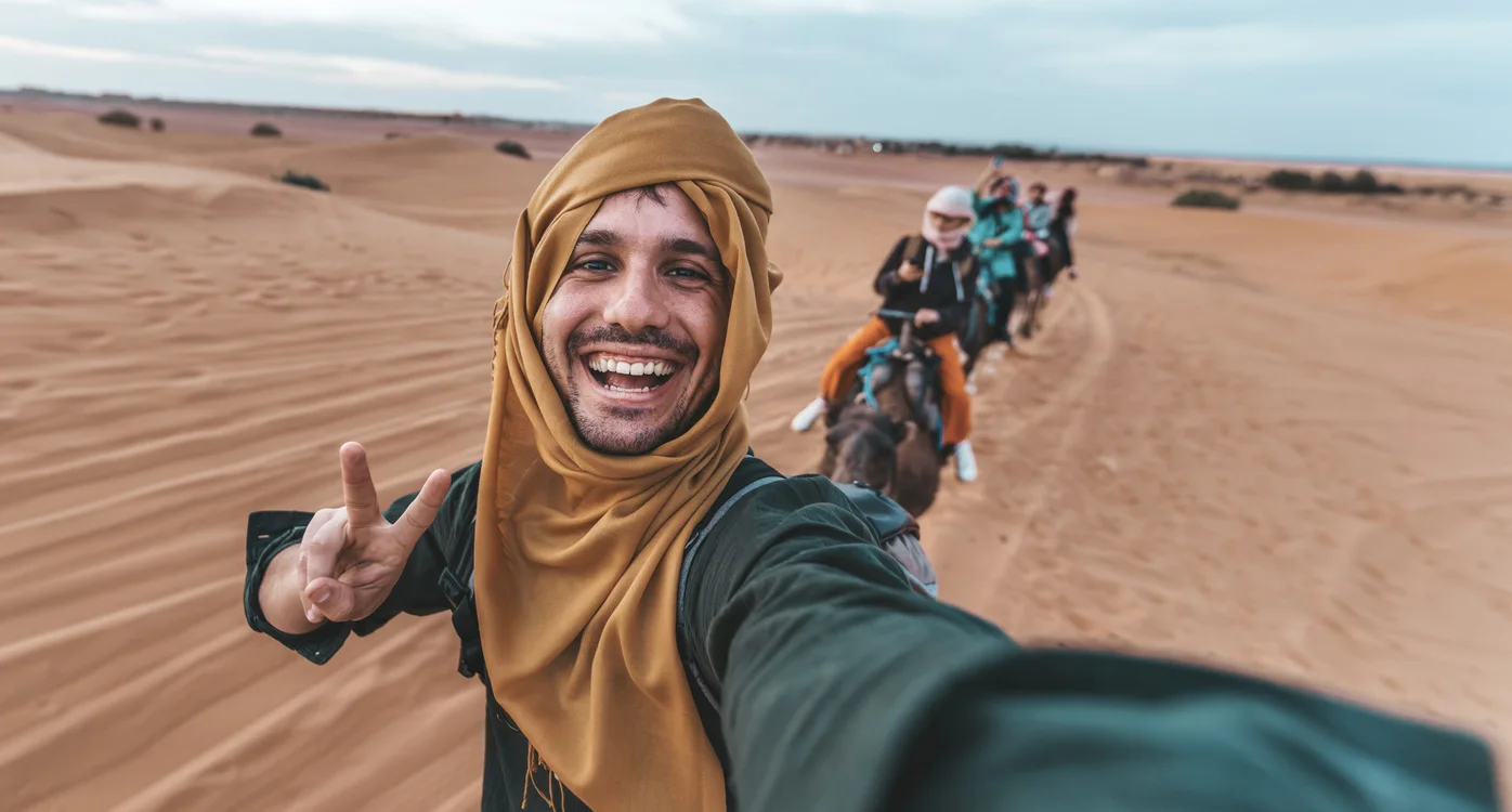 Smiling traveler taking a selfie while riding a camel caravan across the golden desert dunes in Egypt.