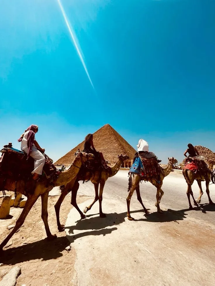 Camel riders approaching the Great Pyramid of Giza under a bright blue sky with a visible sunbeam, capturing a traditional desert scene in Egypt.