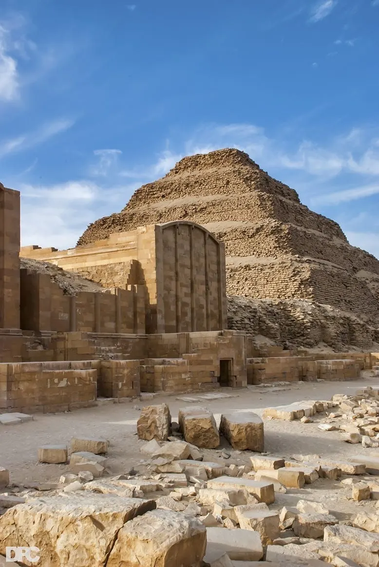 View of the Step Pyramid of Djoser in Saqqara, Egypt, with surrounding ancient stone structures and scattered ruins under a clear blue sky.