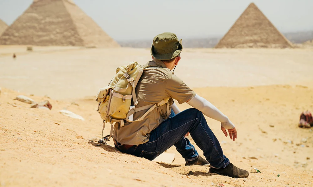 Person sitzt auf einer Sanddüne und blickt auf zwei Pyramiden