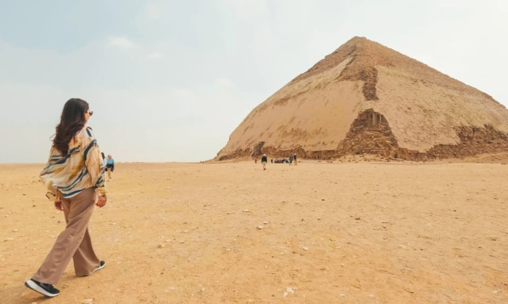 Eine Frau geht von hinten gesehen durch eine weite, sandige Wüstenlandschaft auf die Knickpyramide von Dahschur zu, die die rechte Bildseite dominiert.