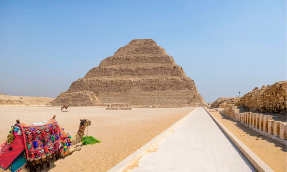 Ein ruhendes Kamel mit bunt gemustertem, rot-blau-grün-gelbem Sattel liegt auf sandigem Boden im Vordergrund einer weiten Wüstenlandschaft; dahinter erhebt sich die mehrstufige Stufenpyramide von Djoser aus hellem Stein unter klarem, blauem Himmel.