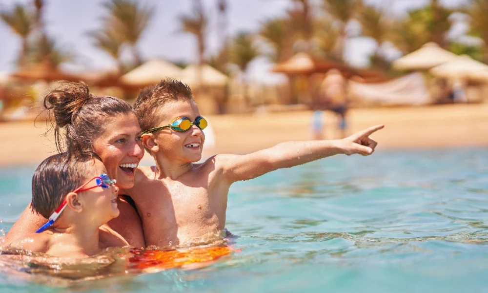 Eine lächelnde Frau und zwei Jungen mit Schwimmbrillen planschen im klaren, türkisfarbenen Wasser eines Strandes; alle blicken nach rechts, wobei der ältere Junge begeistert mit dem Finger in diese Richtung zeigt.