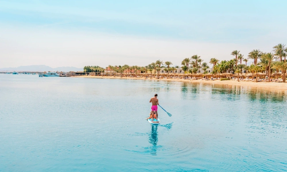 Eine lächelnde Frau und zwei Jungen mit Schwimmbrillen planschen im klaren, türkisfarbenen Wasser eines Strandes; alle blicken nach rechts, wobei der ältere Junge begeistert mit dem Finger in diese Richtung zeigt.