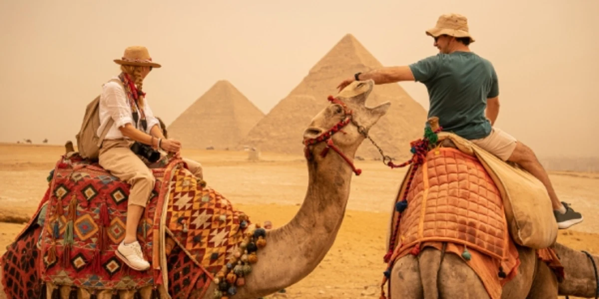 Two tourists riding camels near the Pyramids of Giza, sharing a friendly gesture in the desert haze.