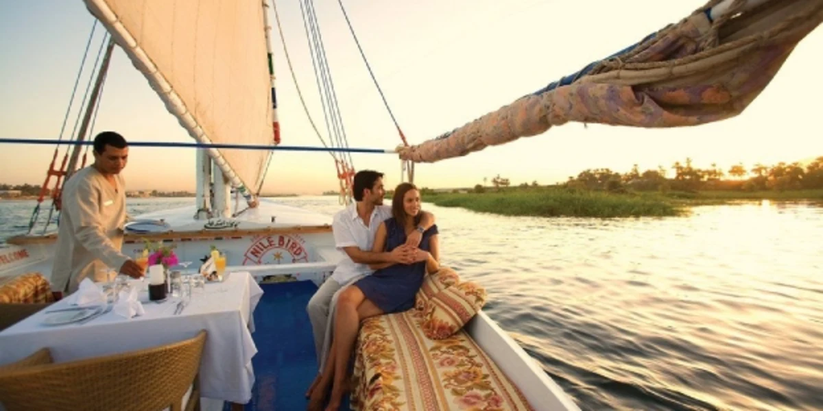 Couple relaxing on a sailboat at sunset, with a server setting a romantic table on calm waters.