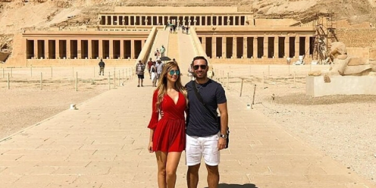 Two tourists standing on the stone pathway leading to Hatshepsut’s Temple, framed by desert cliffs and colonnaded terraces.
