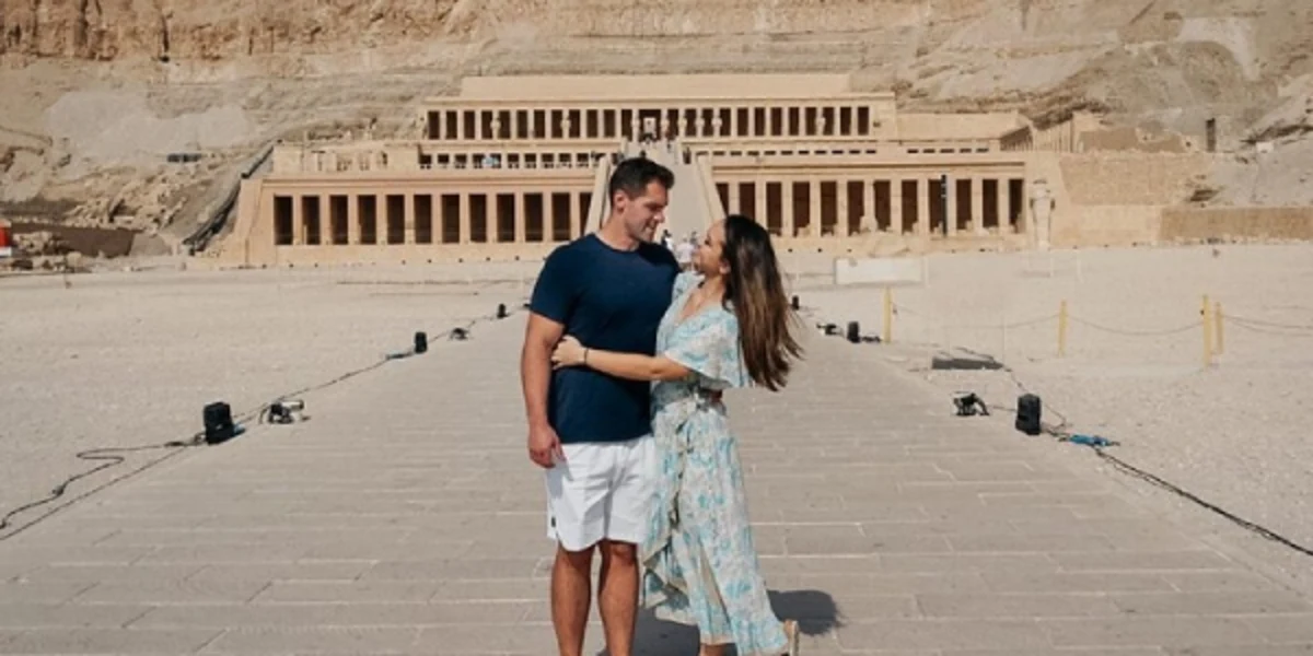 Two visitors stand on a stone path before the Mortuary Temple of Hatshepsut, framed by desert cliffs and colonnaded terraces.