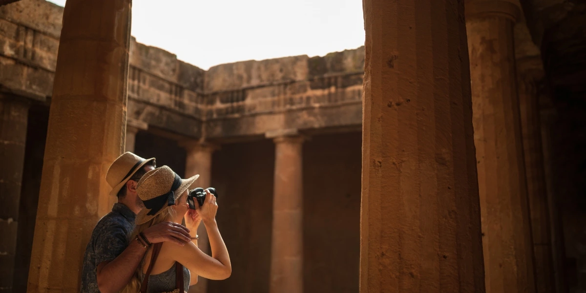 Two tourists exploring an ancient Egyptian temple, one taking a photo amid sunlit stone columns and weathered walls.