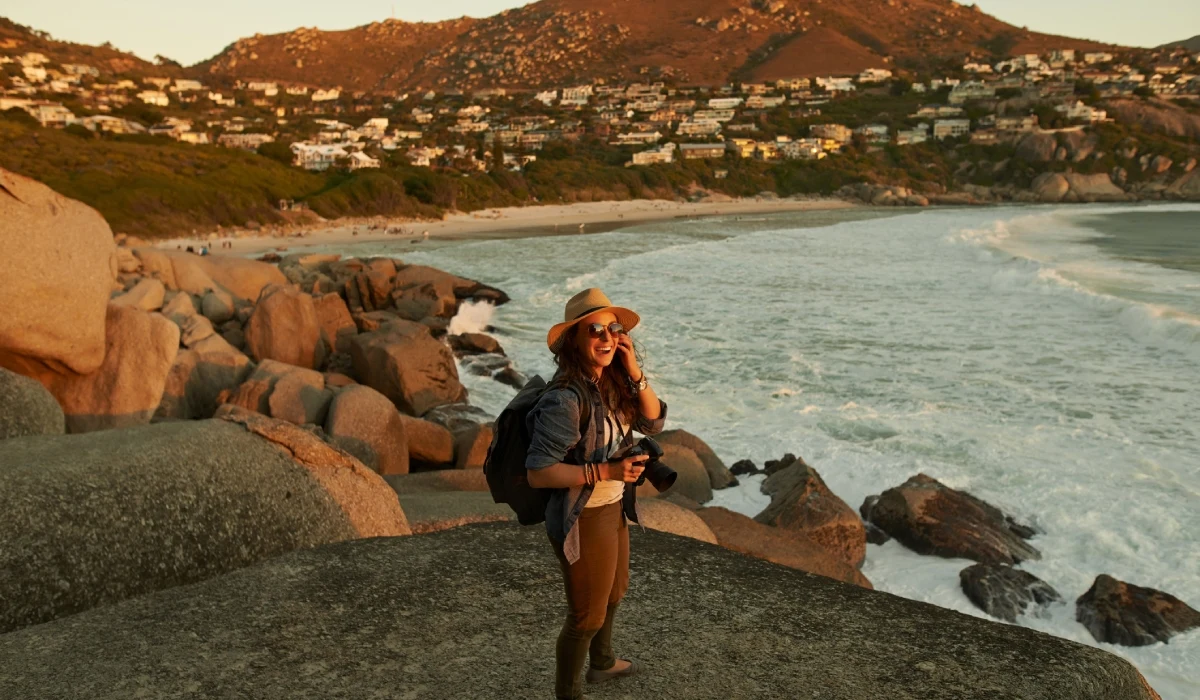 Traveler standing on seaside rock, waves crashing below, overlooking coastal town at sunset.