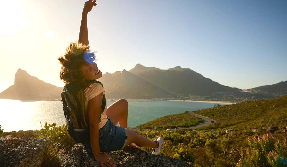 Traveler sitting on rock, arm raised joyfully, overlooking coastal landscape with mountains and sea.