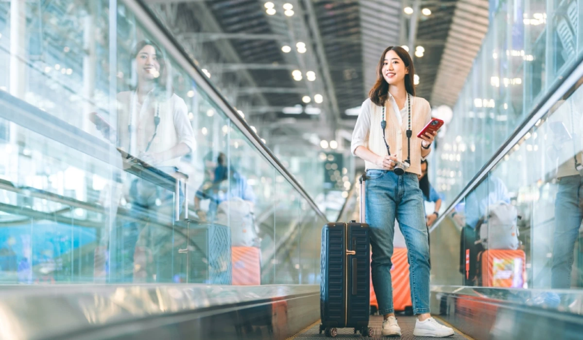 Traveler on a moving walkway in an airport, holding a passport and smartphone with a camera around the neck and a large suitcase beside them.