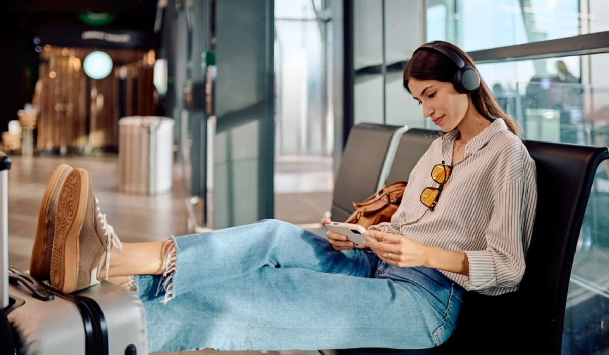  Traveler sitting in an airport terminal with legs resting on a suitcase, wearing headphones and looking at a smartphone, with a café visible in the background.