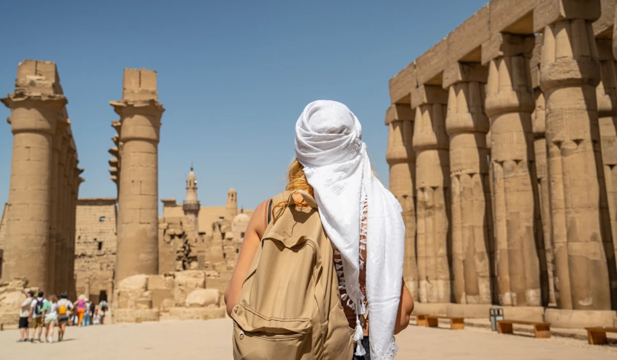 Traveler with a tan backpack and headscarf standing among ancient Egyptian ruins with large stone columns and hieroglyphic carvings, under a clear blue sky.