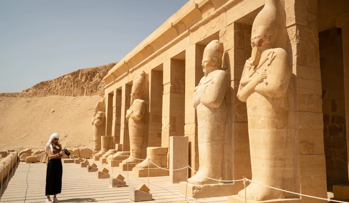 Person standing in front of large sandstone statues at the Mortuary Temple of Hatshepsut in Egypt, with columns and desert cliffs in the background under a clear sky.