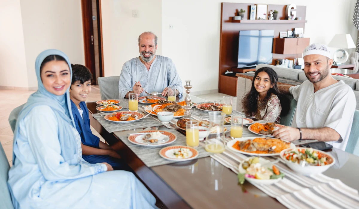  A smiling Moroccan family sits around a large dining table enjoying a traditional meal together. The table is spread with various dishes, including rice, meat, and orange juice, in a brightly lit, modern home setting.