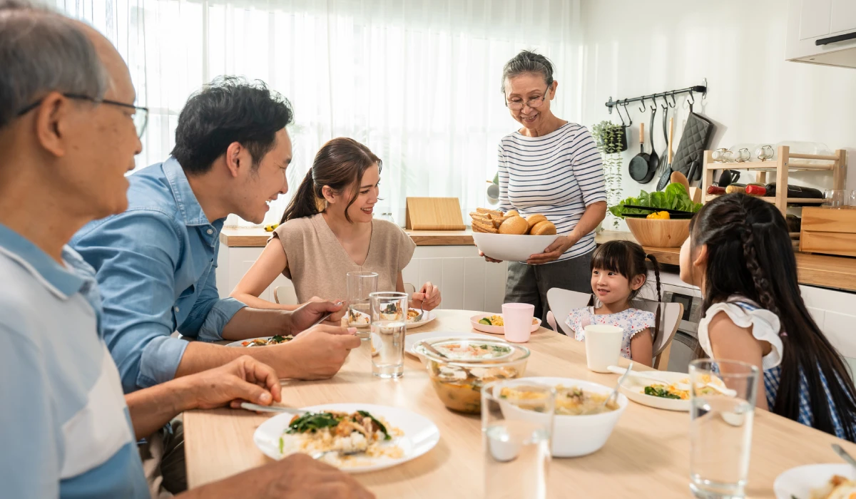A Japanese Family shares a meal in a bright, airy kitchen. An older woman stands and serves a bowl of bread rolls to the group, while children and adults sit around the light wood table filled with various dishes and bowls of soup.