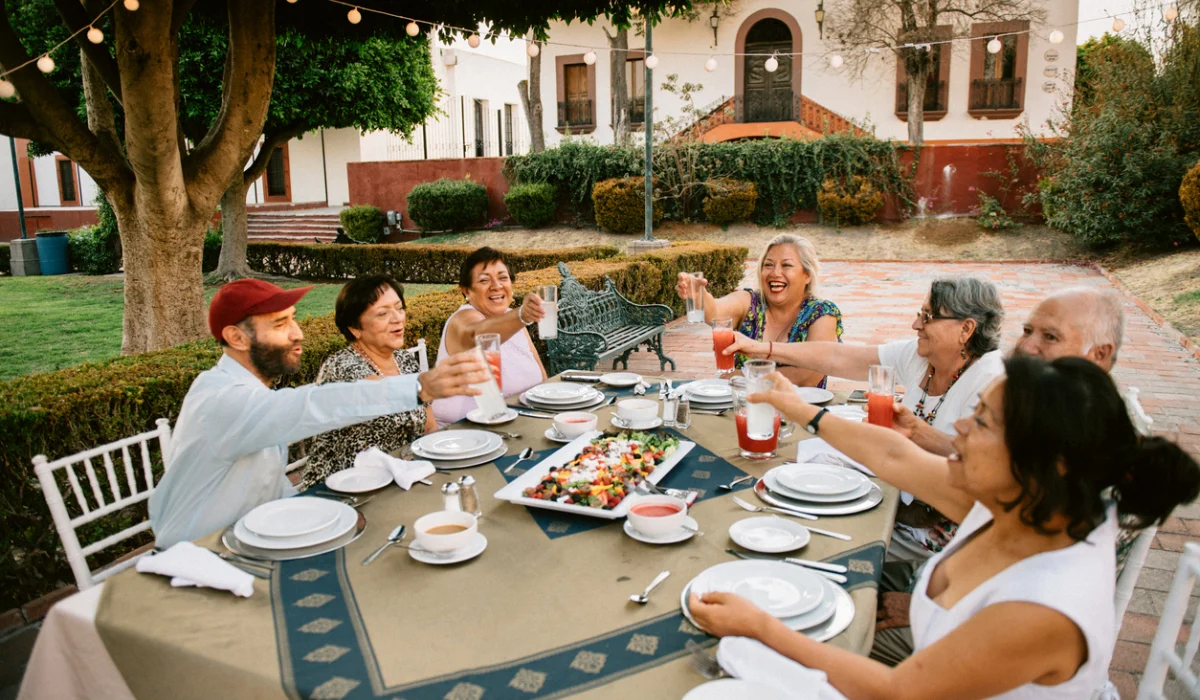 A Mexican Family sits around a dining table in a beautiful outdoor courtyard. The table is elegantly set with white plates and a colorful salad centerpiece, while warm string lights hang from a large tree over the celebratory scene.
