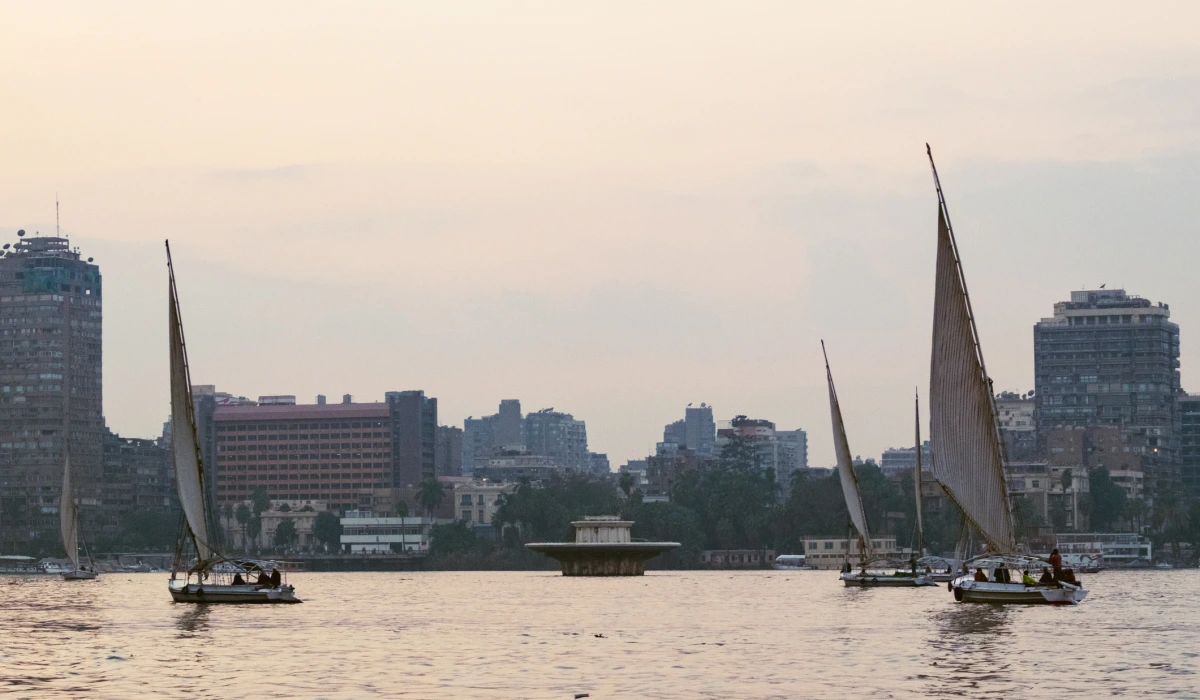 A traditional felucca sailing on the Nile River in Cairo. The boat has a large sail and is surrounded by modern buildings along the riverbank