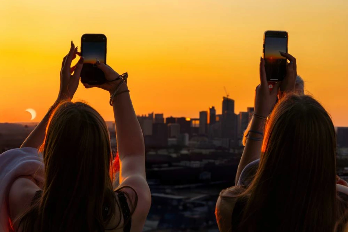 Two travellers photographing the partial phase of a solar eclipse above a city skyline at sunset