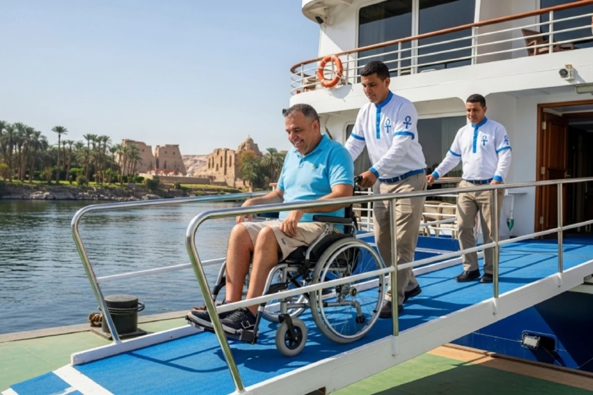   A man in a wheelchair is assisted by two uniformed staff members as he descends a blue ramp from a boat, with palm trees and ancient ruins in the background—showcasing accessible travel in a scenic Egyptian setting.