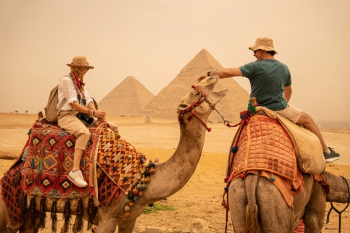 Two tourists riding camels near the Pyramids of Giza, sharing a friendly gesture in the desert haze.