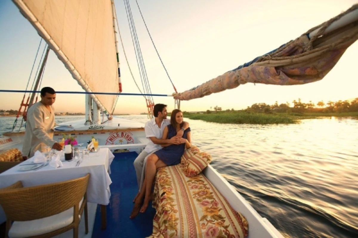 Couple relaxing on a sailboat at sunset, with a server setting a romantic table on calm waters.