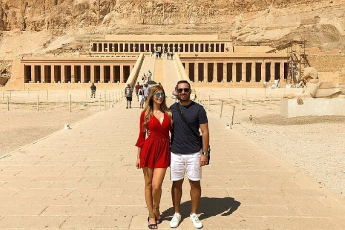 Two tourists standing on the stone pathway leading to Hatshepsut’s Temple, framed by desert cliffs and colonnaded terraces.