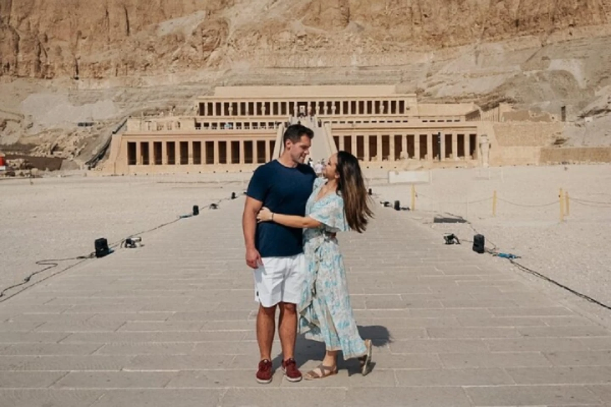 Two visitors stand on a stone path before the Mortuary Temple of Hatshepsut, framed by desert cliffs and colonnaded terraces.