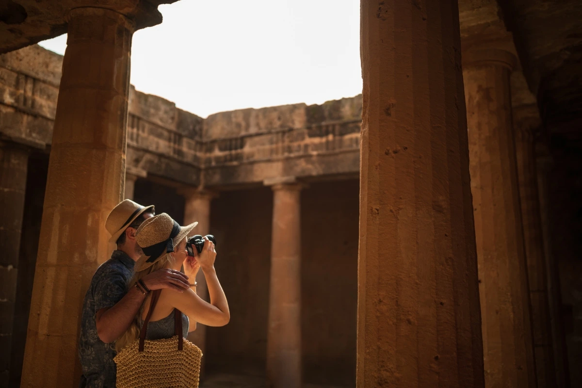 Two tourists exploring an ancient Egyptian temple, one taking a photo amid sunlit stone columns and weathered walls.