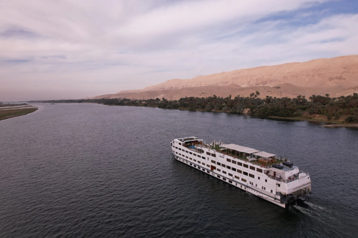 White river cruise ship sailing on a wide river, with lush palm-lined banks and desert hills in the background.