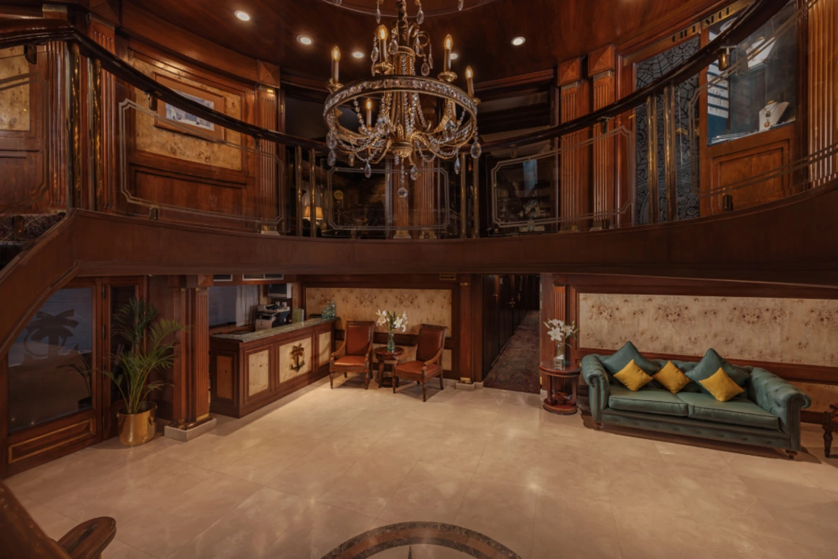 Elegant cruise lobby with a grand chandelier, wood-paneled walls, ornate balcony, and plush seating areas featuring red chairs and a teal tufted sofa.