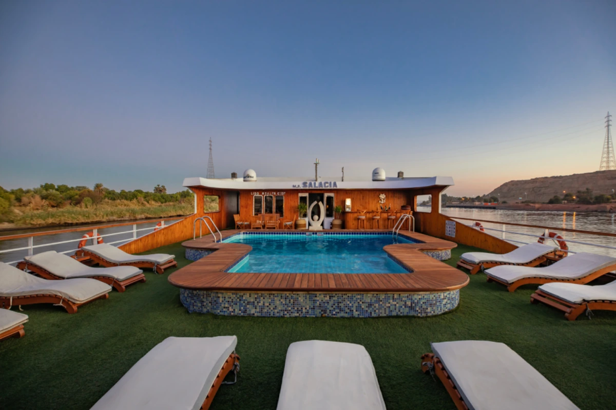 Upper deck of river cruise ship M.S. SALACIA at sunset, featuring a central pool, lounge chairs, and scenic views of greenery and rocky hills.