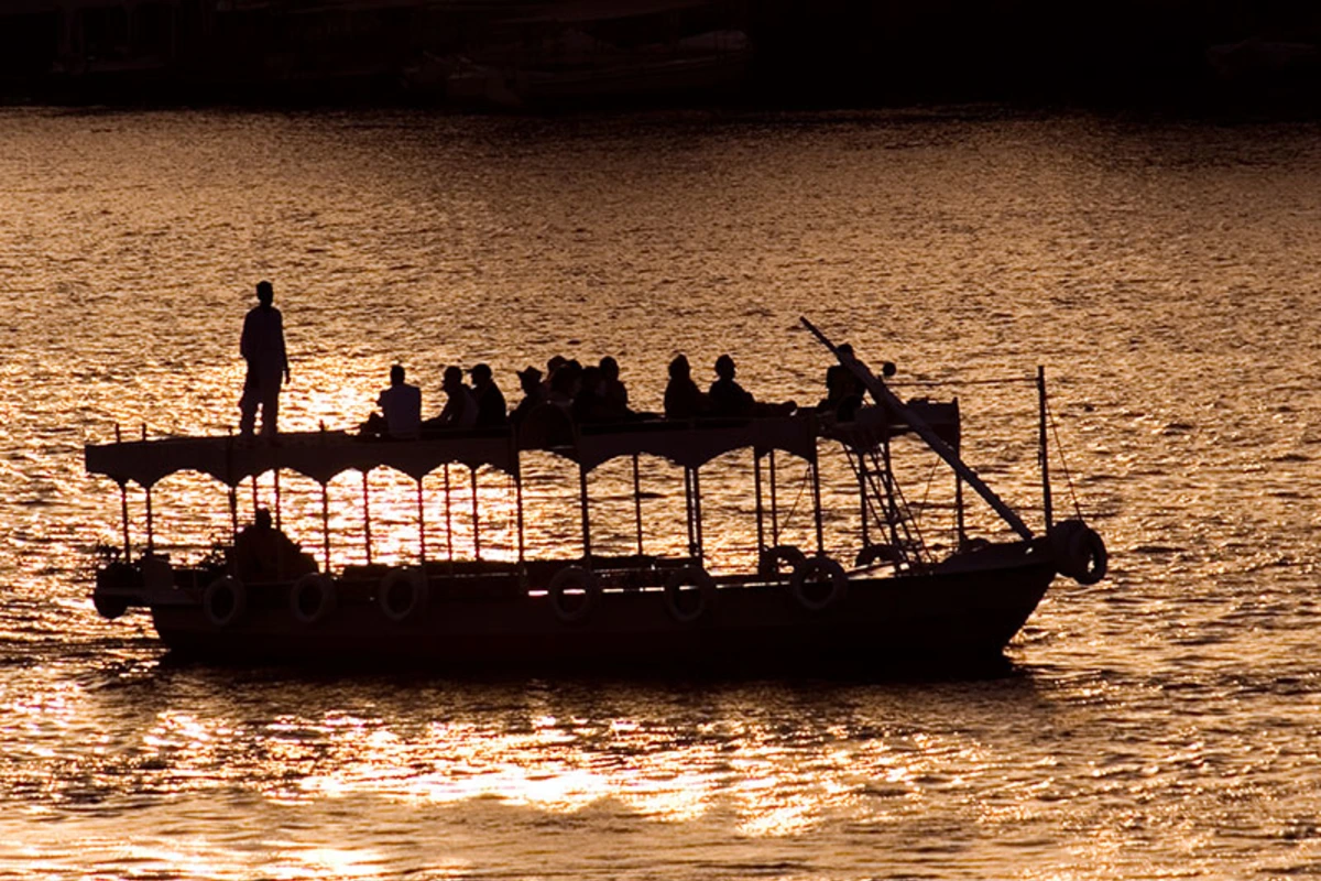 Silhouette eines vollbesetzten Ausflugsboots mit Überdachung auf golden glitzerndem Wasser bei Sonnenuntergang, eine Person steht vorn am Bug.