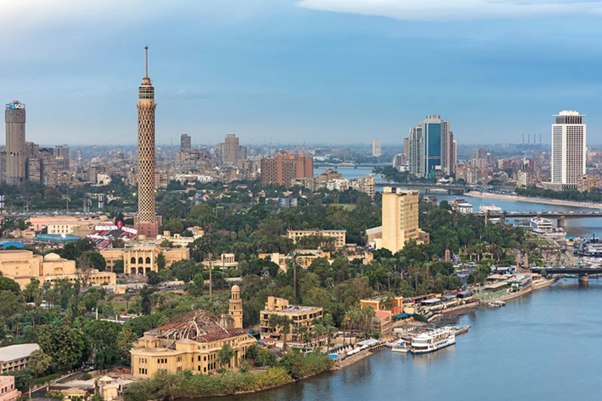 Luftaufnahme von Kairo mit dem Kairoer Turm, Nil, Brücken, historischen Gebäuden mit grünen Kuppeln, Moschee mit Minarett und moderner Skyline unter leicht bewölktem Himmel.