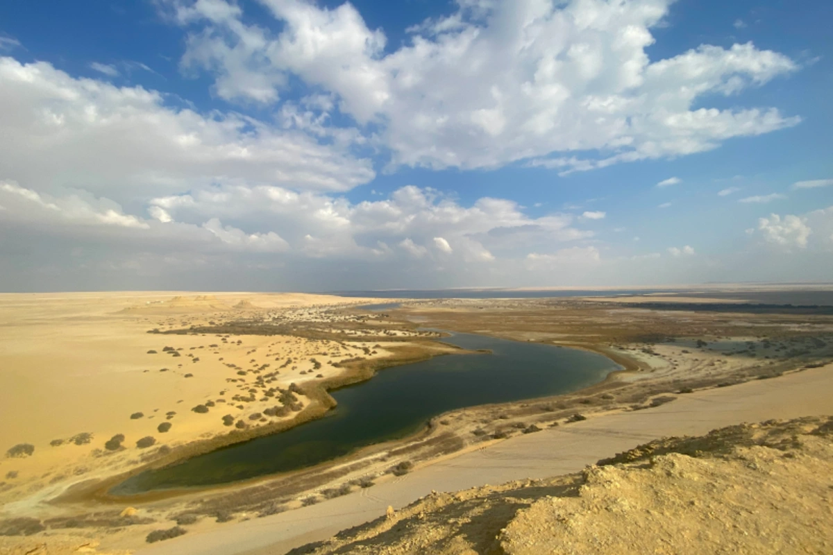Ein atemberaubender Panoramablick auf die Wüste El Faiyum mit einer natürlichen Oase mit ruhigem Wasser, umgeben von goldenen Sanddünen und einem strahlend blauen Himmel.