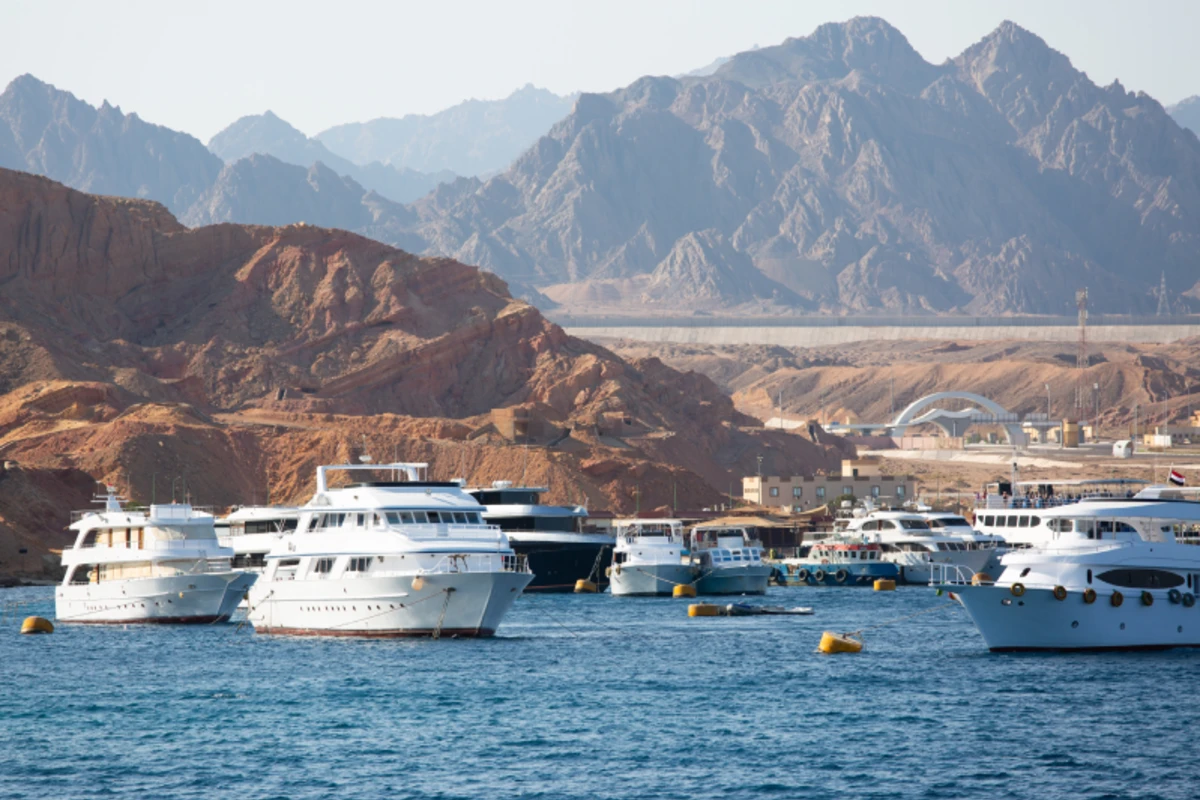 Weiße Yachten und Boote liegen in einer ruhigen Bucht vor einer felsigen Küste mit blauen Bergen im Hintergrund.