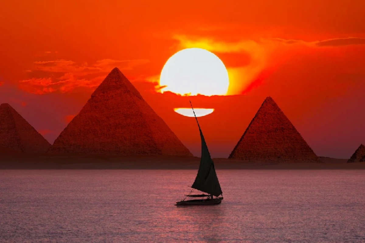 Sonnenuntergang über den Pyramiden von Gizeh: leuchtend roter Himmel, Pyramiden als Silhouetten am Horizont und eine Feluccia mit dunklem Segel auf spiegelndem Wasser im Vordergrund.