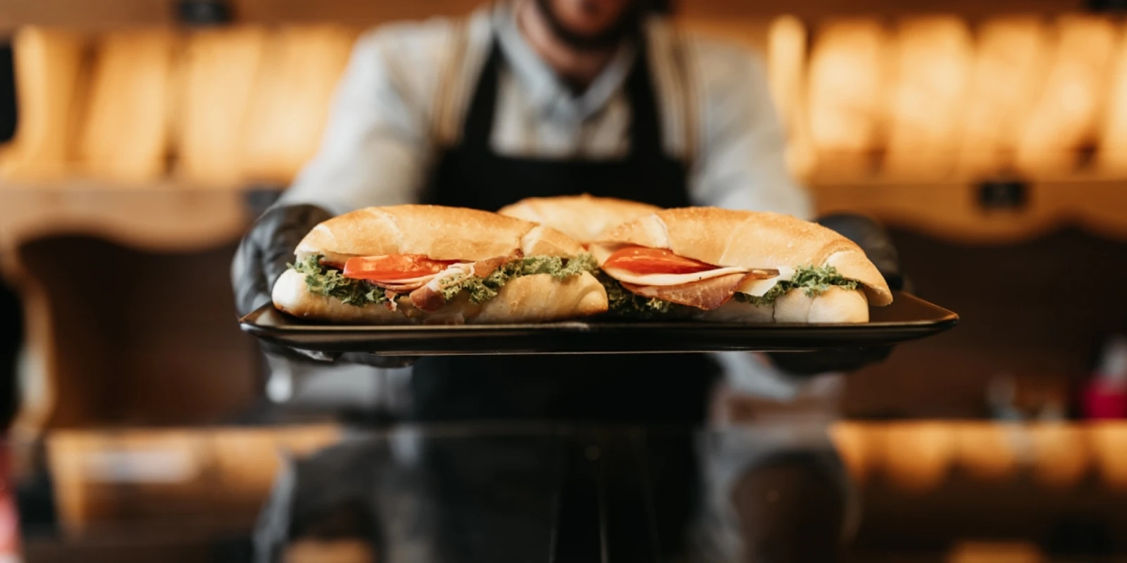 Person in apron holding tray with two fresh sandwiches in a bakery setting.