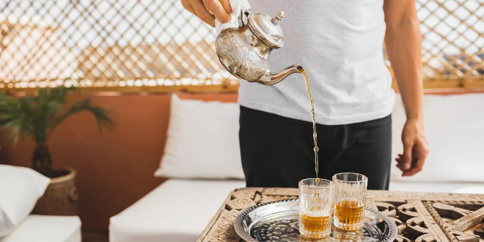 Person pouring golden-brown tea from an ornate silver teapot into two clear glasses on a decorative tray in a cozy lounge setting.