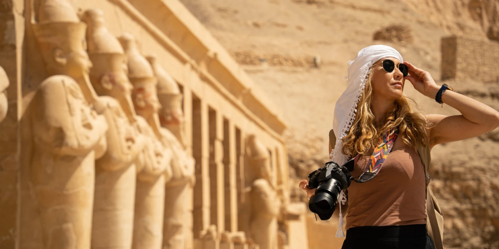 Traveler standing in front of the Mortuary Temple of Hatshepsut in Egypt, wearing sunglasses and a white head covering, holding a camera with colorful strap against a backdrop of sandstone statues and desert cliffs.