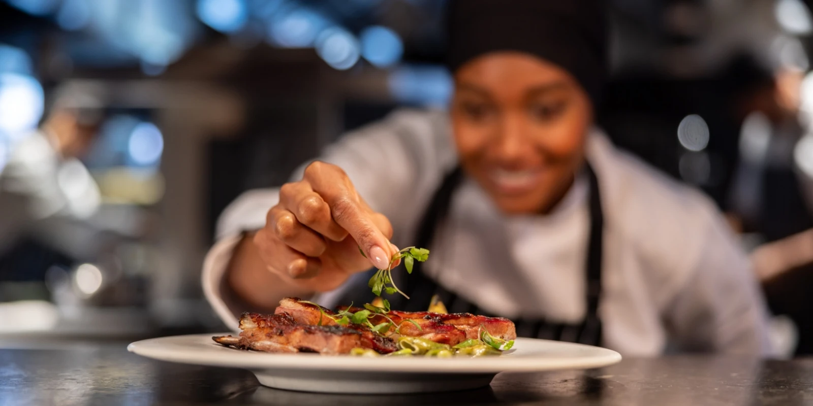  Chef in a professional kitchen carefully placing fresh herbs on a plated dish of cooked meat, with blurred staff and equipment in the background.